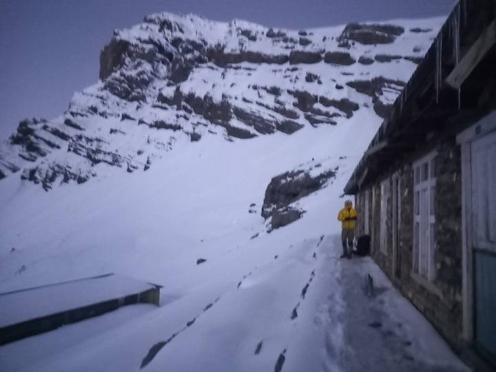 Grainy dusk shot of trekker in yellow jacket outside stone lodge with snowy cliffs behind.