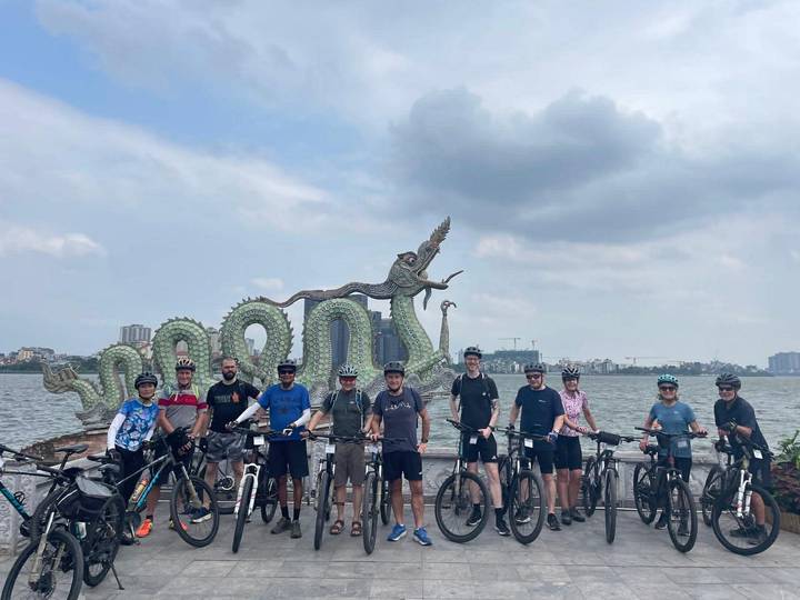Cycling group lines up with bikes in front of dragon sculpture beside large lake in Hanoi.