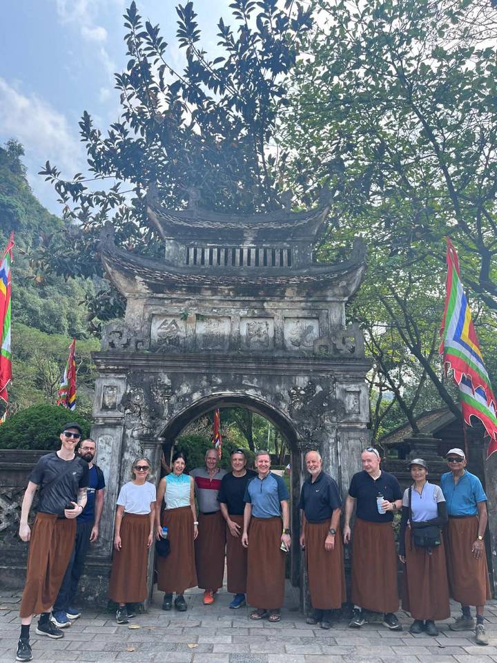 Cycling group poses beneath ancient stone gate adorned with colorful flags and lush trees.