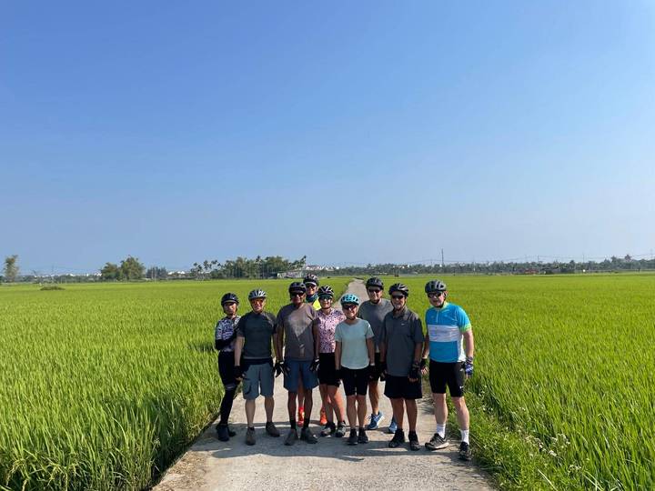 Cyclists stand on narrow rural path surrounded by vibrant green rice paddies under clear sky.
