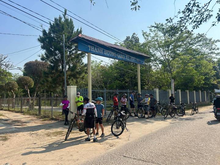 Cyclists rest with bikes beside roadside gate sign in shaded village area.