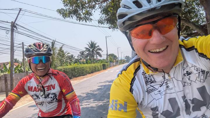 Selfie of two smiling cyclists riding down a rural Vietnamese road lined with palm trees.