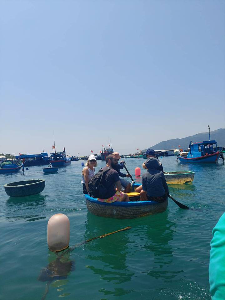 Travelers paddling a small round boat on a calm bay dotted with Vietnamese fishing vessels.