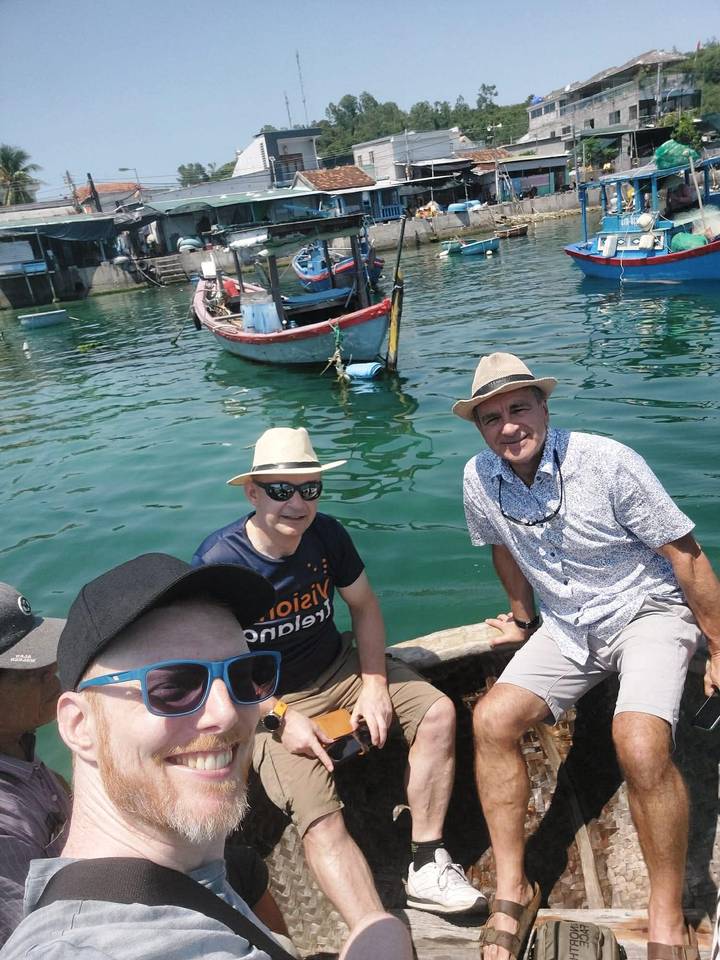 Selfie of three men wearing hats inside a small basket boat on emerald water with fishing boats nearby.