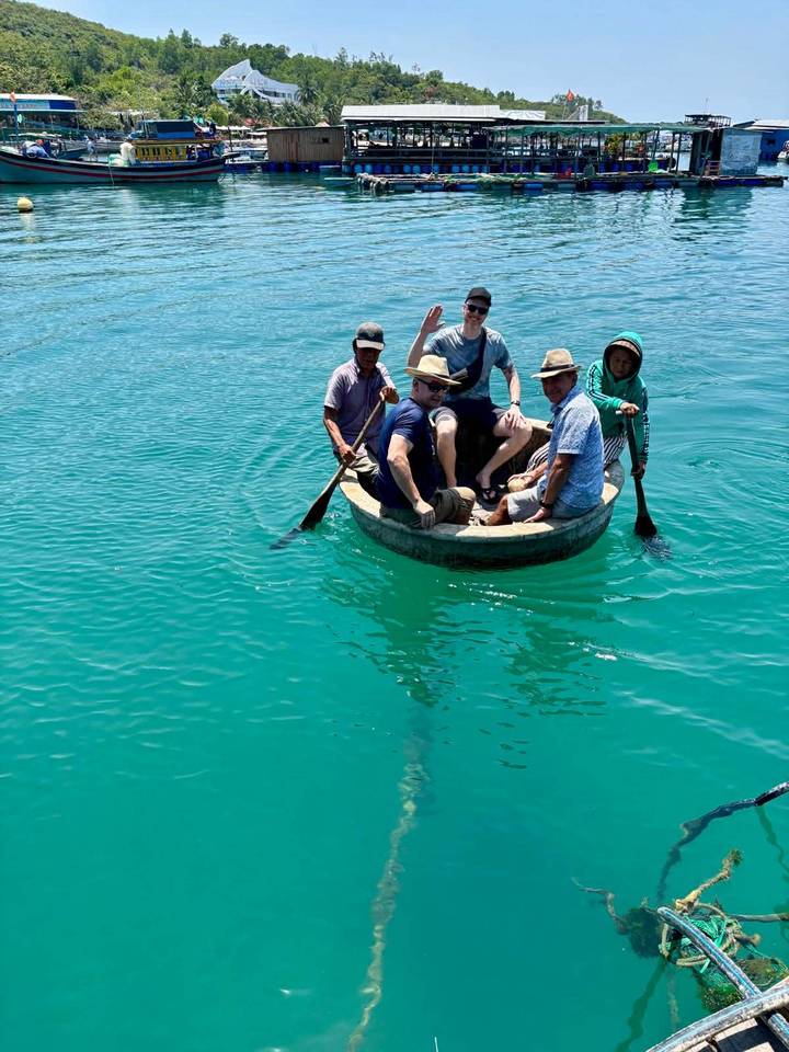 Five travelers smiling and paddling a round basket boat on bright turquoise water.