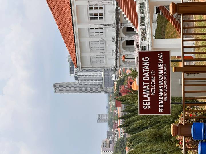 Sign welcoming visitors to the Melaka Museum Corporation with city skyline behind.