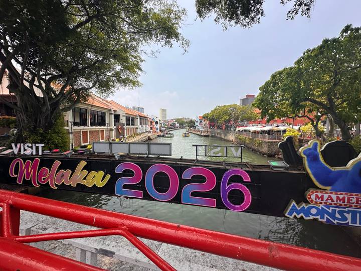 Colorful sign 'Visit Melaka 2026' spanning a pedestrian bridge over the Melaka River.