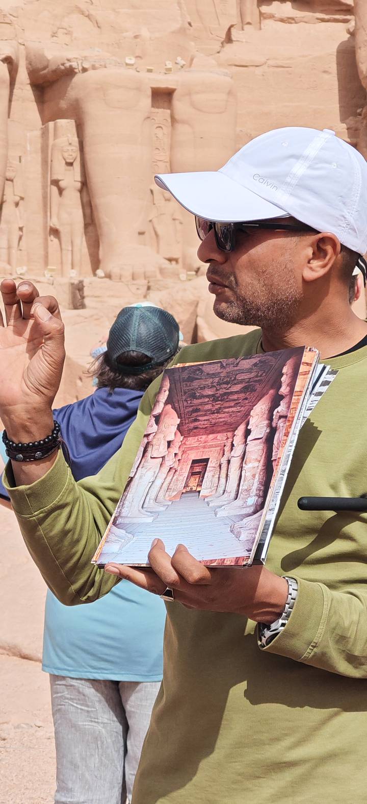 Tour guide holds a photo book depicting Abu Simbel temple while explaining to visitors amid sandstone ruins.