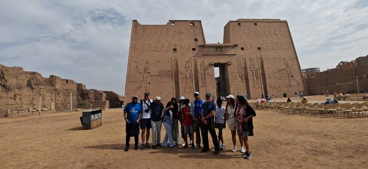 Tour group poses in front of the monumental sandstone pylons of Edfu Temple under a bright sky.