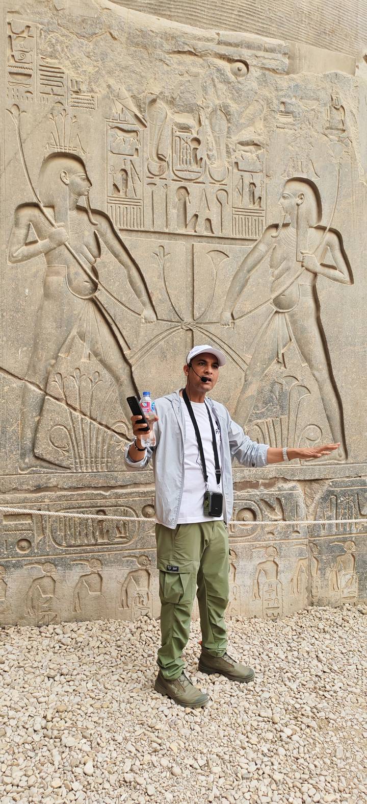 Egyptian guide speaking through headset microphone in front of carved hieroglyphic stone reliefs.