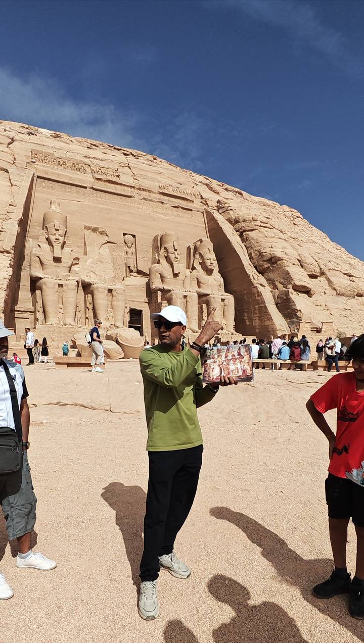 Guide shows illustration while pointing at the colossal seated statues of Abu Simbel temple surrounded by visitors.