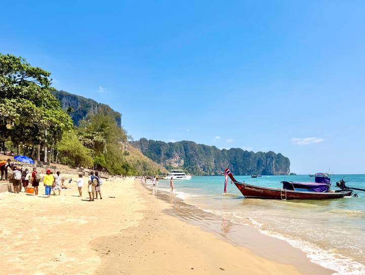 Tropical sandy beach in Krabi with long-tail boat anchored in turquoise water, limestone cliffs in the background and visitors strolling along the shore.