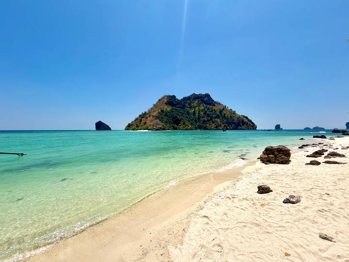 Secluded island rising from crystal-clear turquoise waters bordered by a white sandy beach and scattered rocks.