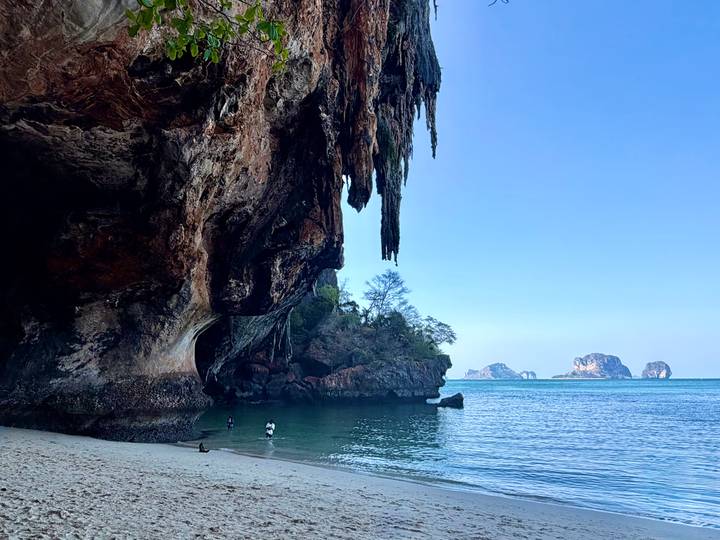 Sheer limestone overhang framing a quiet cove with shallow emerald water; a couple of swimmers explore the beach.