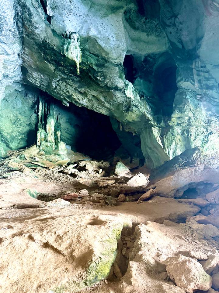 Dimly lit limestone cave interior with stalactites, green mineral stains and rocky floor.