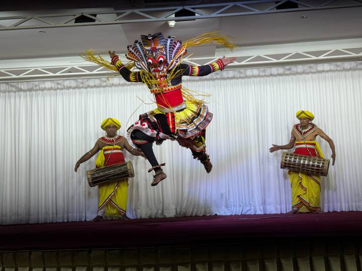 Energetic traditional Kandyan dancer mid-air with vibrant costumes accompanied by two drummers on stage.