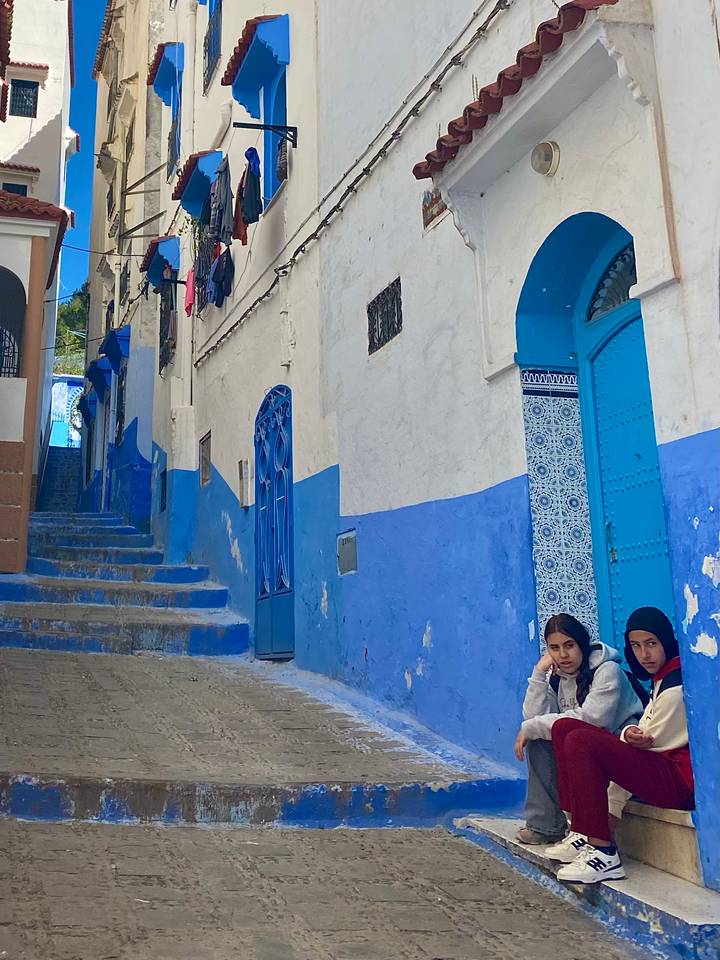 Blue-painted alley in Chefchaouen with patterned tiled doorway and two local girls seated on the step.