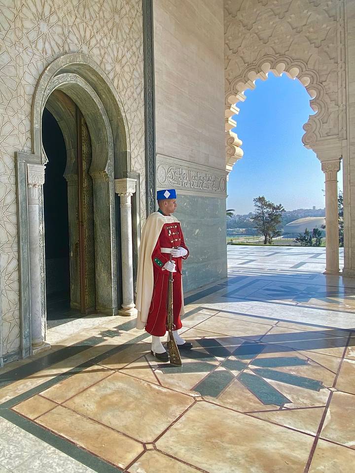 Uniformed royal guard in bright red attire stands at the marble entrance of the Mausoleum of Mohammed V with cityscape beyond.