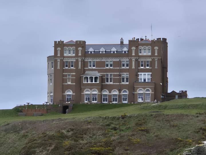 Large brown stone building resembling a coastal castle sits on a grassy hill under overcast skies.