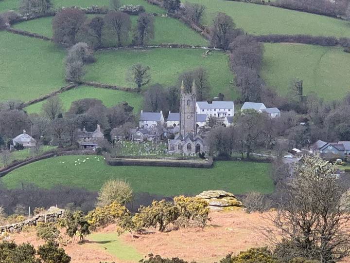 Distant view of a small English village with stone church and fields framed by rolling green hills.