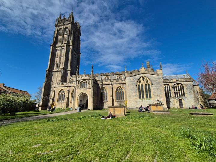 Tall Gothic church tower and nave set against a bright blue sky with visitors relaxing on the lawn.