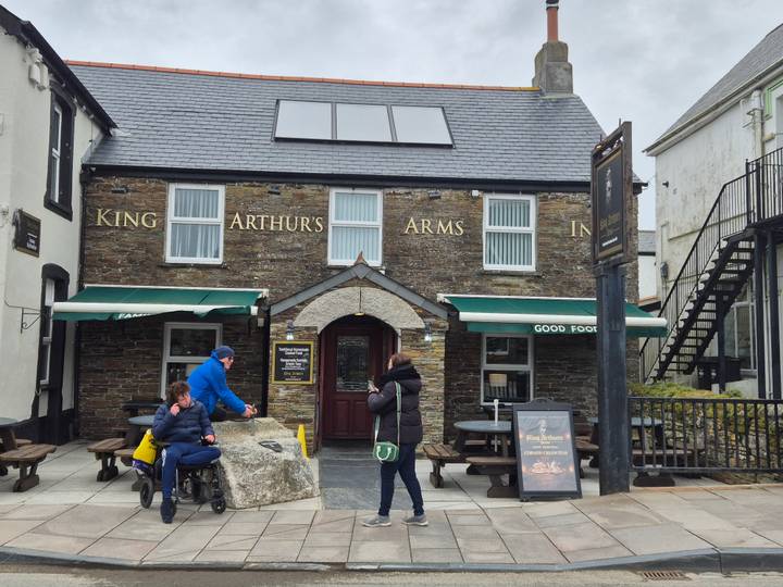 Stone-fronted King Arthur’s Arms Inn with green awnings as visitors read the menu outside.