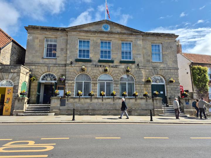 Historic stone Town Hall with hanging flower baskets while pedestrians walk past on a sunny day.