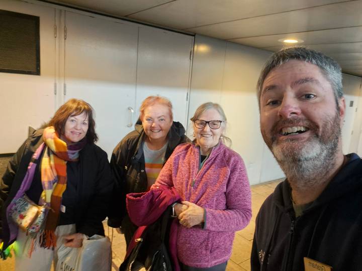 Group selfie of four cheerful travellers in an indoor corridor with bright lights overhead.