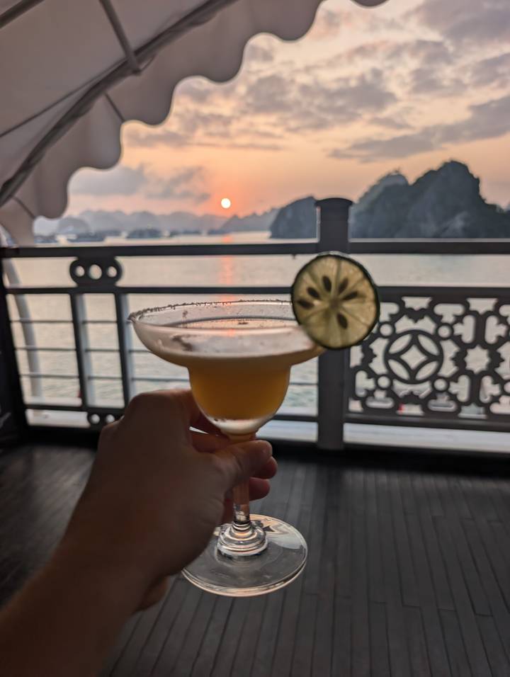 Hand holding a cocktail at sunset aboard a boat with limestone islands silhouetted in Halong Bay