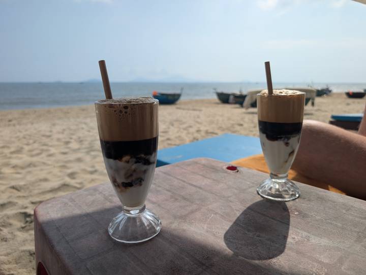 Two tall iced coffees set on a rustic beach table overlooking a calm sandy shoreline