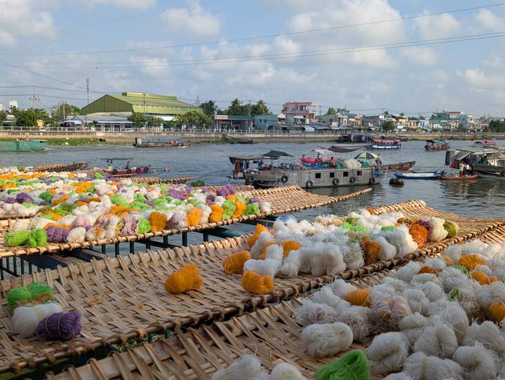 Bundles of colourful rice noodles drying on woven racks beside a bustling Mekong river port