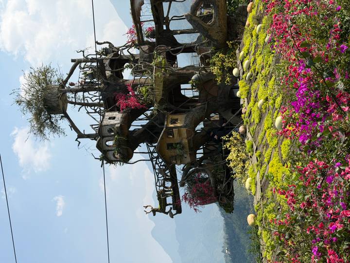 Whimsical multi-level treehouse structure surrounded by lush flowers and mountain backdrop.
