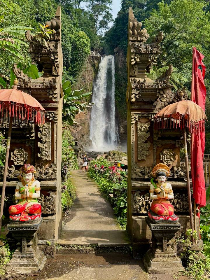 Tall waterfall framed by ornate Balinese temple gate and colorful tropical plants.