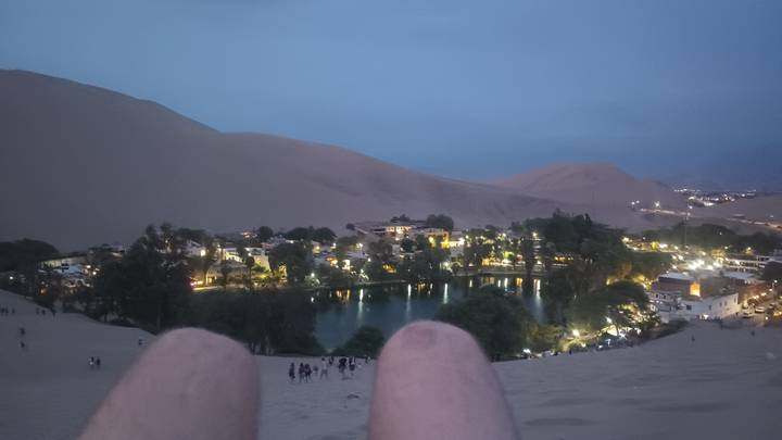 Evening view over a desert oasis village with lit palm-lined lagoon, seen from a sand dune with traveller’s knees in foreground.