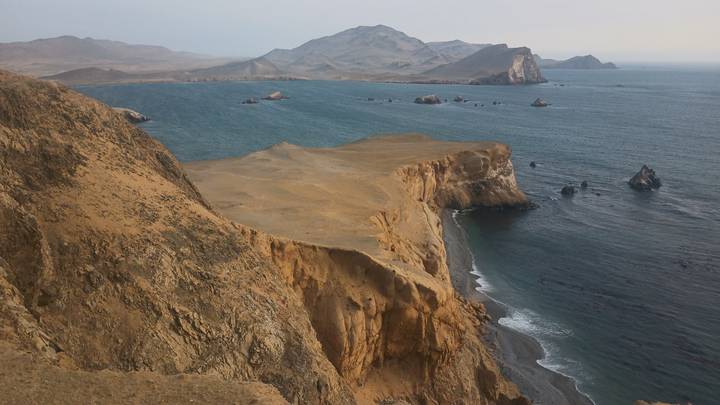Wide coastal panorama of rugged tan headlands meeting deep blue ocean with scattered rock stacks.