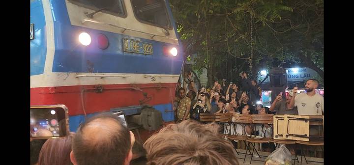 Night scene of a train passing inches away from packed café tables while tourists film the spectacle.