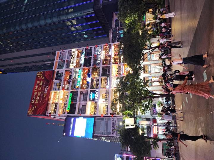Bustling pedestrian street at dusk in front of a high-rise filled with illuminated cafés and shops.