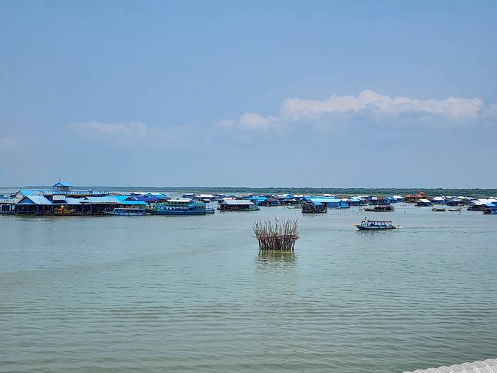 Floating village of blue-roofed houses spread across a vast lake on a clear day.