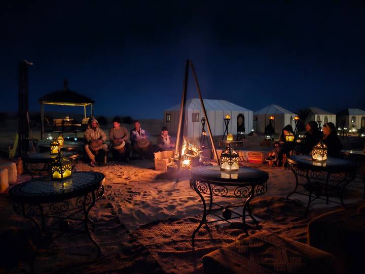 Travellers gather around a campfire with drums and lanterns at a desert camp.