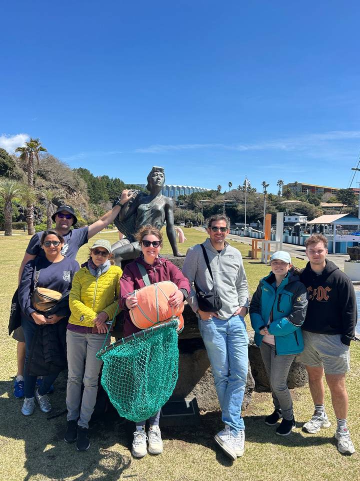 Travellers pose by a seaside mermaid statue on a sunny, breezy day.