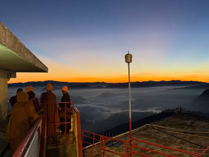 Monks in saffron robes watch a colorful dawn over mist-filled valleys from a mountain summit railing.