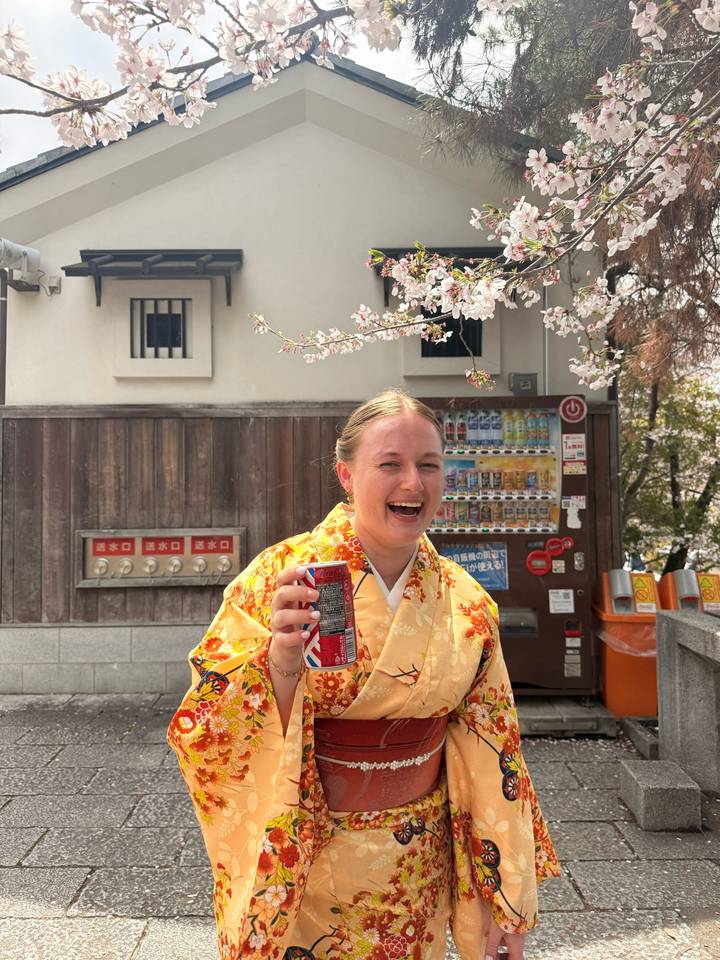 Young woman in a yellow kimono laughing beneath cherry blossoms while holding a drink.