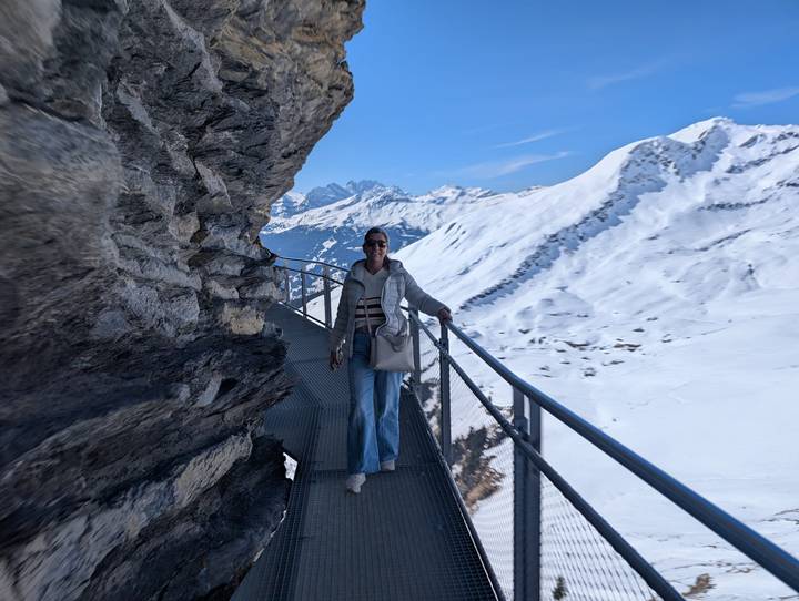 Woman standing on a metal cliff walkway overlooking snow-covered alpine peaks.