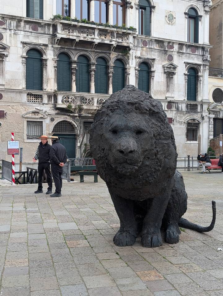 Textured black lion sculpture in a Venetian square with two gondoliers chatting nearby.