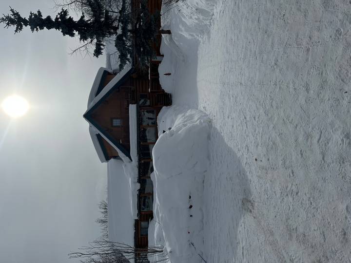 Snow-covered log lodge under a bright hazy sun with thick drifts surrounding the entrance.