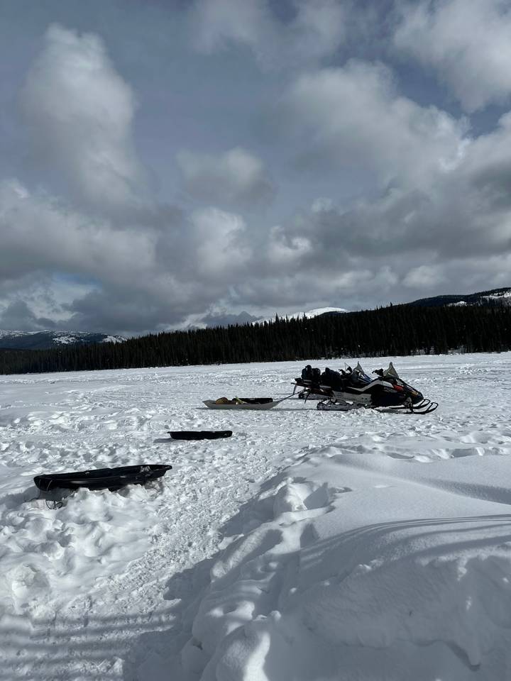 Snowmobiles and sleds parked on a vast frozen lake surrounded by dark evergreen forests.