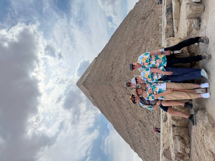 Matching-shirt group posing cheerfully in front of a massive pyramid under dramatic skies.