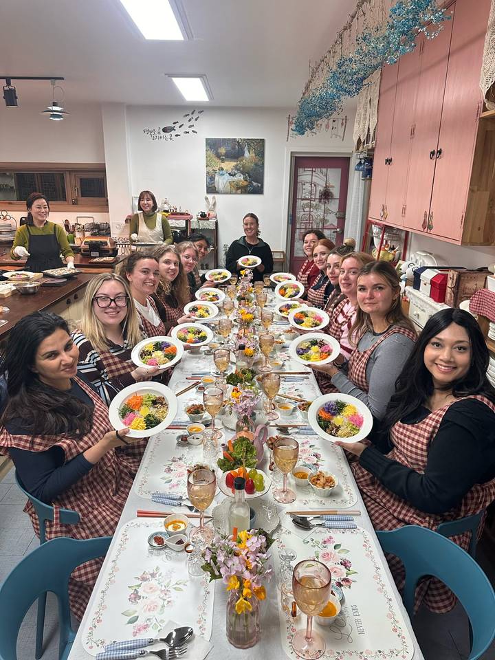 Large group of travellers wearing checked aprons sit at a long table proudly holding colourful rice bowls in a cooking studio.