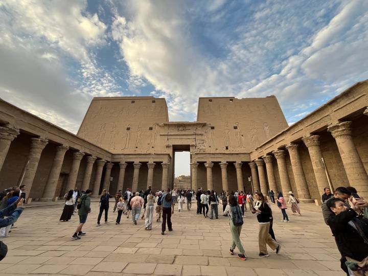 Wide courtyard of an ancient Egyptian temple lined by tall columns and bas-reliefs under a partly cloudy sky, filled with visitors.