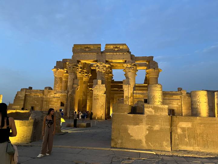 Golden lighting highlights the columns and carvings of Kom Ombo Temple at dusk as visitors wander around.